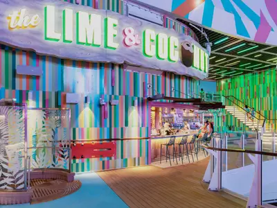 Innenansicht eines farbenfrohen Bereichs auf der Utopia of the Seas. Eine Bar mit Gästen und Barkeepern ist sichtbar, darüber ein leuchtendes Schild 'the LIME & COCONUT'. Eine breite Treppe und eine Duschkabine mit Blattmuster sind ebenfalls erkennbar. Die Wände sind in bunten vertikalen Streifen gestaltet, was eine lebendige Atmosphäre schafft.