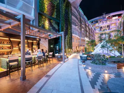 Central Park auf der Utopia of the Seas bei Nacht: Beleuchtete Trellis Bar, vertikale Gärten, Parkanlage. Im Hintergrund Balkonkabinen und Wasserrutschen derselben.