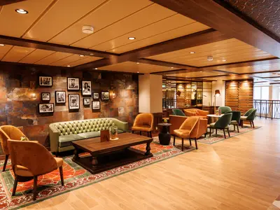 Gemütlicher Lounge-Bereich an Bord der Utopia of the Seas. Eine rustikale Wand mit Schwarz-Weiß-Fotos, ein grünes Sofa und orangefarbene Sessel laden zum Verweilen ein. Der Bereich ist mit einem gemusterten Teppich und Holzmöbeln ausgestattet.