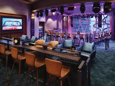 Die Music Hall der Utopia of the Seas zeigt eine beleuchtete Bühne, diverse Sitzbereiche mit Barhockern und Sesseln sowie eine Fensterfront. Links ist ein Bildschirm zu sehen.