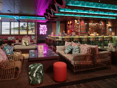 Tropische Bar auf der Utopia of the Seas. Mit Tresen, Barhockern, Tiki-Dekos und Sitzbereichen. Pinkes Neonlicht dominiert, ergänzt durch ein Strandfenster.