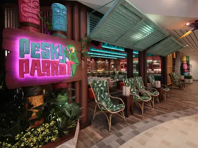 Die tropische "Pesky Parrot" Bar an Bord der Utopia of the Seas, mit leuchtendem Schild, Tiki-Figuren und Rattan-Sitzgruppen.