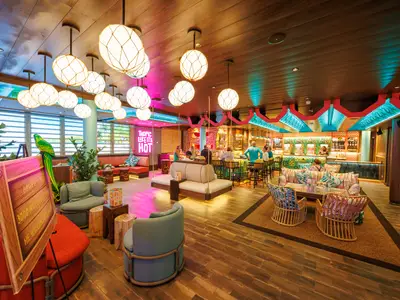 Eine lebhafte Lounge an Bord der Utopia of the Seas mit einer bunten Bar, bequemen Sitzgelegenheiten, tropischen Pflanzen und stimmungsvoller Beleuchtung.