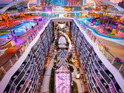 Ein hoher Blick auf die Utopia of the Seas zeigt die zentrale, begrünte Promenade, gesäumt von Balkonkabinen, sowie lebendige Pool- und Wasserparkbereiche auf den Oberdecks.