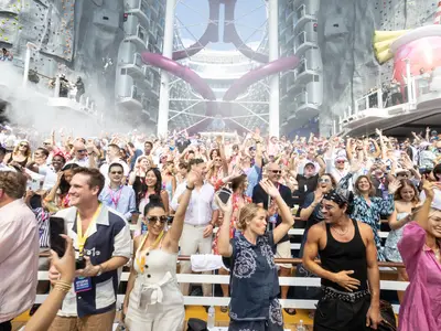 Jubelnde Passagiere auf den Außendecks der Utopia of the Seas. Im Hintergrund sind eine Kletterwand, lila Wasserrutschen und eine Cocktail-Skulptur bei einer Deck-Show zu sehen.