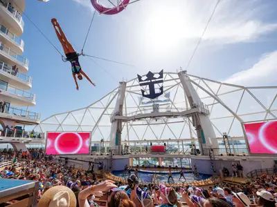 Eine Akrobatin hängt kopfüber über dem AquaTheater-Pool auf der Utopia of the Seas. Zuschauer sehen Wassershow mit Darstellern, LED-Wänden und Royal Caribbean Logo.