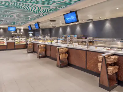 Innenansicht eines modernen Selbstbedienungsrestaurants auf der Utopia of the Seas. Sichtbar sind mehrere Buffetauslagen, digitale Anzeigetafeln, eine grüne Blattmuster-Decke und Holzverkleidungen.