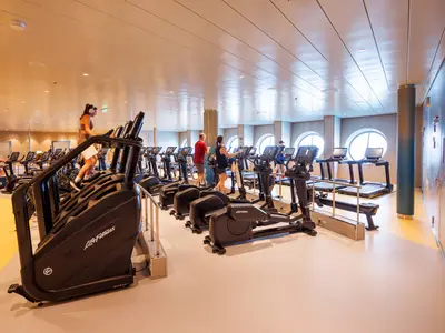 Blick in ein modernes Fitnessstudio auf der Utopia of the Seas mit mehreren Personen an Laufbändern, Ellipsentrainern und Stair Climbern vor runden Fenstern.