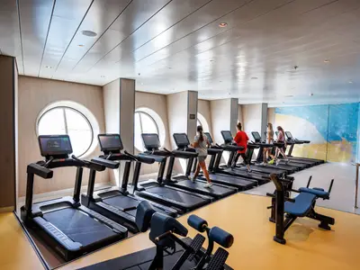 Das Fitnessstudio der Utopia of the Seas. Personen nutzen Laufbänder, zu sehen sind runde Fenster, eine blaue Wandmalerei und Hantelbänke auf gelbem Boden.