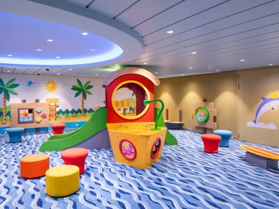 Farbenfroher Kinderspielbereich an Bord der Utopia of the Seas mit Rutsche, interaktiven Wandspielen und bunten Sitzhockern auf einem Wellen-Teppich.