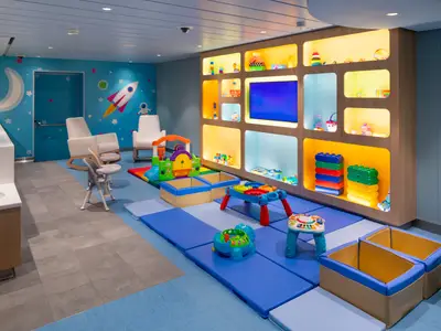 Ein heller Kinderspielbereich auf der Utopia of the Seas. Man sieht eine blau gestrichene Wand mit Weltraum-Motiven, weiße Schaukelstühle, ein Regal mit Spielzeug, gepolsterte blaue Spielmatten und bunte Spielgeräte auf dem Boden. Links befindet sich ein Bereich mit Waschbecken und Ablagefläche.