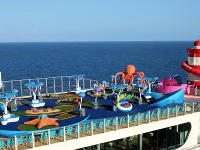 Auf dem Deck der Utopia of the Seas ist ein maritimer Minigolfplatz zu sehen, verziert mit einem riesigen orangefarbenen Oktopus und einem Leuchtturm mit Wasserrutsche. Im Hintergrund erstreckt sich das Meer.