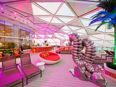Ein weitläufiger, futuristisch anmutender Lounge-Bereich auf der Utopia of the Seas, beleuchtet in Magenta-Tönen. Man sieht eine Bar, diverse Sitzmöbel, eine silberne Skulptur und Pflanzen unter einem Glasdach.