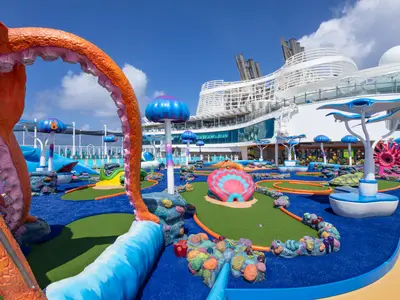 Bunter Mini-Golf-Platz mit Unterwasser-Thema auf dem Deck der Utopia of the Seas. Fantasievolle Korallen, Muscheln und Pilzformen auf blauem und grünem Kunstrasen, mit dem Schiff im Hintergrund.