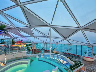 Innenansicht des Solariums der Utopia of the Seas. Helle Pools, Liegen und Meerblick durch große Fenster. Eine geometrische Dachkonstruktion überspannt den Raum.