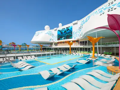Sonniges Pooldeck der Utopia of the Seas mit mehreren Pools, vielen Liegen im und am Wasser sowie einer großen LED-Wand. Bunte Dekorationen sind unter blauem Himmel zu sehen.
