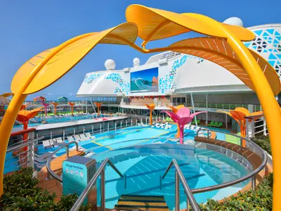 Blick auf das farbenfrohe Pooldeck der Utopia of the Seas. Mehrere Pools, Liegestühle, bunte Schirme und ein großer Bildschirm mit Surfszene.