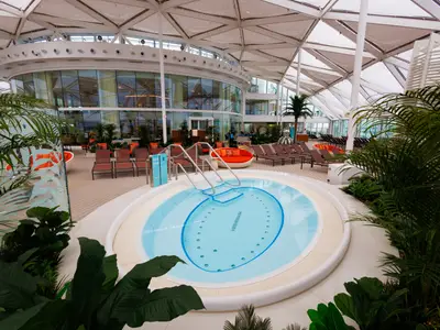 Innenansicht des Solariums auf der Utopia of the Seas: Runder Whirlpool, Liegestühle, Palmen und ein verglastes Gebäude unter einem hellen Dach.
