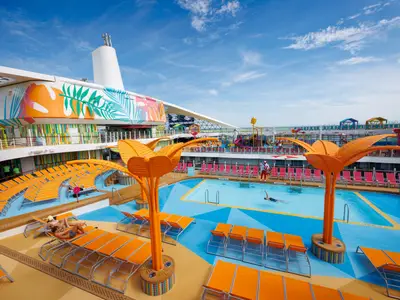 Ansicht des Pooldecks der Utopia of the Seas: Schwimmbecken, orange Liegen, Deko-Palmen, Wasserpark, Bademeister. Blauer Himmel.