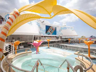 Das Pooldeck der Utopia of the Seas zeigt Pools, einen Whirlpool und bunte Schatten spendende Strukturen. Ein großer Bildschirm ist sichtbar.