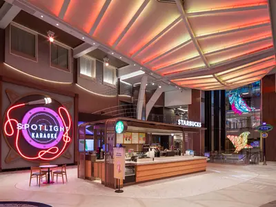Innenansicht der Utopia of the Seas. Ein Starbucks-Kiosk, ein Neon-Schild 'Spotlight Karaoke', farbige Deckenbeleuchtung und große, beleuchtete Fisch-Kunstinstallationen.