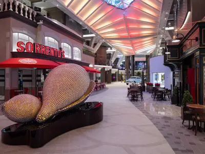 Die helle, thematisierte Promenade der Utopia of the Seas zeigt eine Pizzeria, eine große Zitrus-Skulptur und einen Pub. Im Hintergrund ist ein Oldtimer zu sehen.