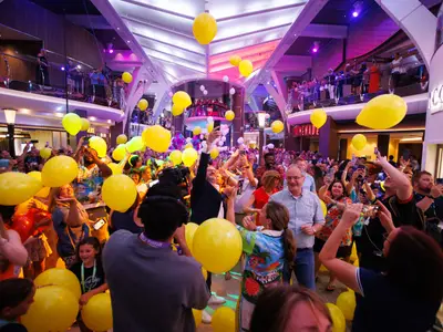 Gelber Ballonregen auf dem Promenadendeck der Utopia of the Seas. Viele Passagiere fangen Ballons und fotografieren das festliche Treiben in der großen Schiffshalle.