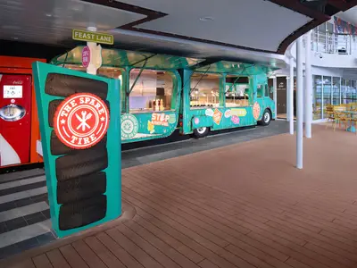 Auf dem Deck der Utopia of the Seas ist ein türkiser Food-Truck-Bereich namens "The Spare Tire" zu sehen, ergänzt durch einen Coca-Cola Automaten und Sitzgelegenheiten im Hintergrund.