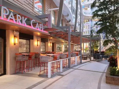 Utopia of the Seas: Abendaufnahme des Park Cafe mit beleuchtetem Schild, Außenterrasse, orangefarbenen Stühlen. Dahinter die Glasarchitektur des Schiffs und begrünte Bereiche.