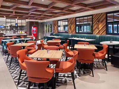 Modernes Restaurant mit orangefarbenen Stühlen, Holztischen und einer langen Sitzbank. Im Hintergrund ein Buffetbereich. Decke und Wände sind mit Holzelementen und Pflanzen gestaltet. Ein roter Getränkeautomat steht seitlich.