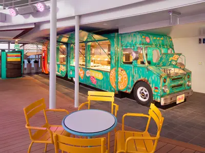 Foodtruck-Restaurant 'The Spare Tire' auf dem Deck der Utopia of the Seas. Der türkisblaue Truck im Vintage-Stil steht bei gelben Tischen und Stühlen. Im Hintergrund ist das Meer.