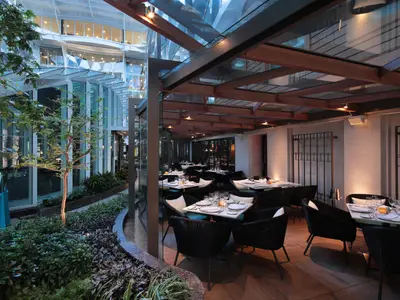 Ein modernes Restaurant mit elegant gedeckten Tischen und dunklen Korbsesseln unter einem Glasdach. Außen ist üppige grüne Vegetation zu sehen, die eine beruhigende Atmosphäre schafft. Das Restaurant ist hell beleuchtet mit warmem Licht.