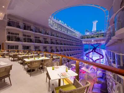 Außenbereich des Giovanni’s Italian Kitchen & Wine Bar auf der Utopia of the Seas am Abend. Zahlreiche Tische und Stühle sind elegant gedeckt. Im Hintergrund sind mehrere Balkonkabinen und die beleuchtete Rutschenanlage sowie das Meer zu sehen.
