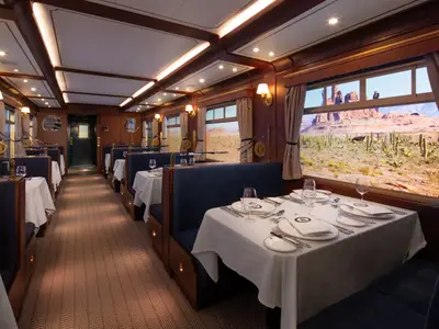 Innenansicht eines eleganten Restaurants im Zug-Stil mit blauen Sitzbänken und weiß gedeckten Tischen. Durch die Fenster ist eine beeindruckende Wüstenlandschaft mit Kakteen und Felsformationen zu sehen.