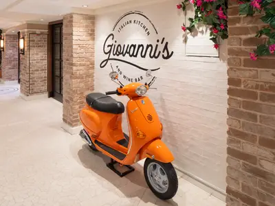 Utopia of the Seas: Orangefarbener Roller vor 'Giovanni's Italian Kitchen'. Rechts Ziegelwand mit rosa Blumen, links Gang. Heller Boden.