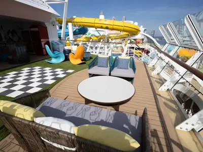Das obere Deck der Utopia of the Seas an einem sonnigen Tag. Es zeigt eine Terrasse mit Holz- und Kunstrasenflächen, bequemen Sitzmöbeln und einem schwarz-weiß karierten Feld. Im Hintergrund sind farbenfrohe Wasserrutschen und weitere Decks zu sehen.