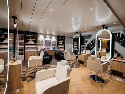Friseursalon auf der Utopia of the Seas. Zu sehen sind Stylingstühle, beleuchtete ovale Spiegel, Waschbecken, Produktregale und eine Wand mit Blättermuster.