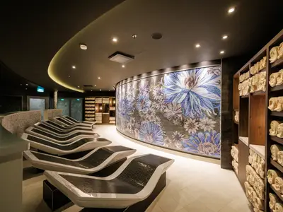 Modernes Spa mit ergonomischen Liegen und blauer Blumenmosaikwand. Rechts offene Regale voller gerollter Handtücher. Indirekte Deckenbeleuchtung sorgt für Ruhe.