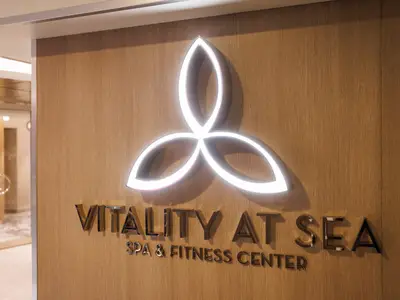 Auf der Utopia of the Seas ist eine Holzwand mit dem beleuchteten, dreiblättrigen Logo und dem Schriftzug 'Vitality at Sea Spa & Fitness Center' zu sehen. Im Hintergrund ist ein Eingangsbereich erkennbar.