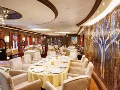 Elegantes Restaurant der Utopia of the Seas. Festlich gedeckte Tische, helle Stühle und eine farbige Mosaikwand mit Baummotiv. Holztäfelung und Weinkeller runden das Ambiente ab.
