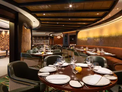 Blick in ein stilvolles Restaurant an Bord der Utopia of the Seas mit gedeckten Tischen, grünen Stühlen und einer kunstvoll beleuchteten Wand.