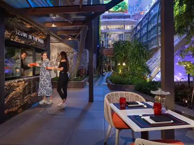 Abendliche Aufnahme auf der Utopia of the Seas zeigt Gäste am Izumi Imbiss in einer begrünten Promenade, daneben sind Esstische zu sehen und im Hintergrund die beleuchteten Decks des Schiffes.