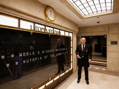 Formal gekleidetes Crewmitglied steht vor digitaler Anzeigetafel mit 'Utopia Station' und 'WORLD'S BIGGEST WEEKEND Utopia OF THE Seas'. Eine goldene Wanduhr ist sichtbar.