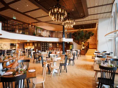 Ein stilvolles, zweistöckiges Restaurant der Utopia of the Seas mit zahlreichen Tischen, einem Olivenbaum und einer Treppe. Helle Fenster und Kronleuchter.