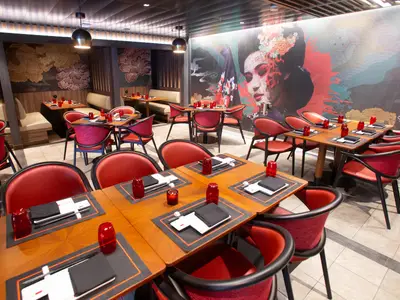 Blick in ein Restaurant auf der Utopia of the Seas. Es ist im asiatischen Stil eingerichtet, mit vielen roten Stühlen, Holztischen und einer auffälligen Wandmalerei. Tischgedecke sind vorbereitet.