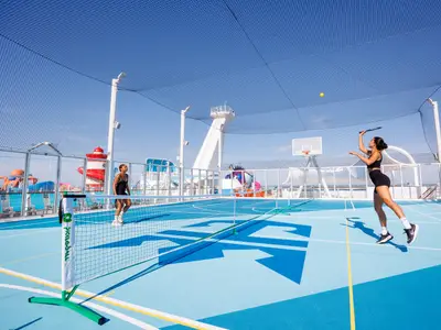 Zwei Frauen spielen auf dem Sportdeck der Utopia of the Seas Pickleball. Eine schlägt den Ball, im Hintergrund Wasserrutschen.