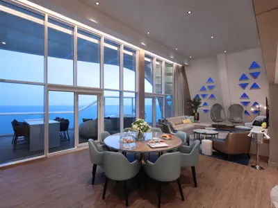 Sicht in eine Suite der Utopia of the Seas: Ess- und Wohnbereich mit raumhohen Fenstern, Meerblick und Balkon mit Whirlpool.