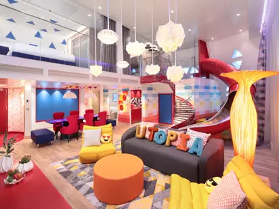 Innenansicht einer bunten, zweistöckigen Suite auf der Utopia of the Seas. Mit Schlafloft, Wohnbereich, Esstisch und einer auffälligen roten Rutsche.