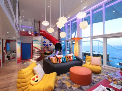 Innenansicht einer farbenfrohen Suite auf der Utopia of the Seas mit roter Rutsche, Wendeltreppe und Meerblick. Bunte Sitzmöbel und farbige Dekoration prägen das helle Interieur.
