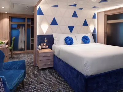 Helle Kabine der Utopia of the Seas. Ein großes Bett mit blauen Kissen und ein geometrisches Kopfteil sind zentral. Blauer Sessel und Kleiderschrank links zu sehen.